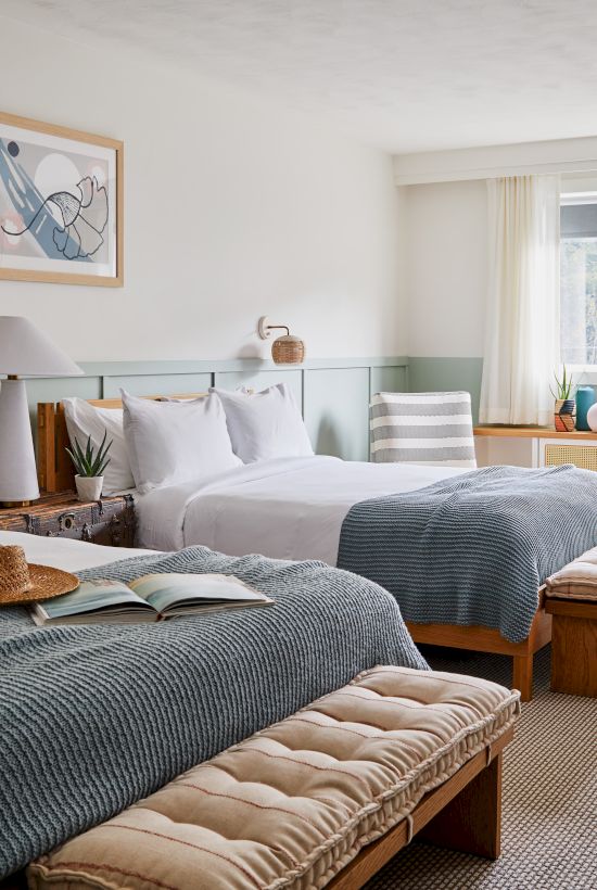 A cozy bedroom with two beds, soft blankets, a hat, a table, chairs, art on the wall, and a large window letting in natural light.