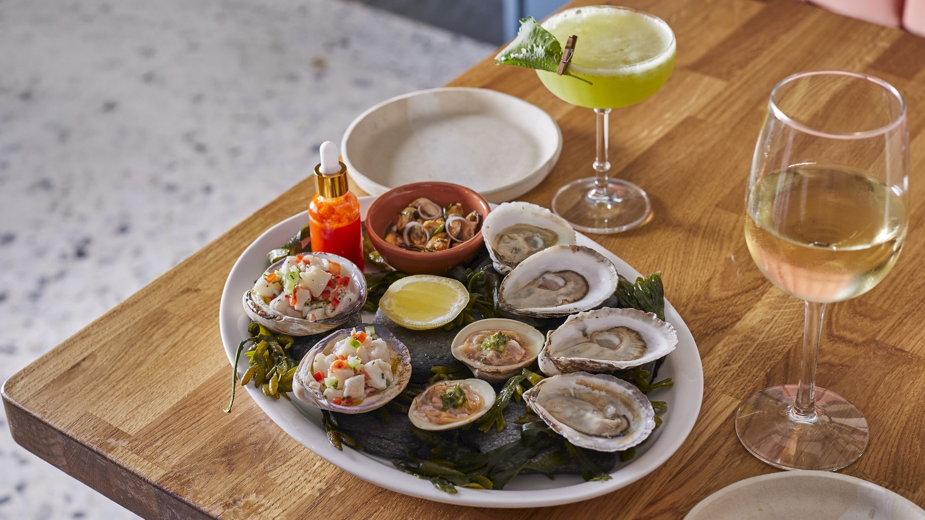 A plate of fresh oysters and clams served with hot sauce and lemon, alongside a green cocktail and a glass of white wine.