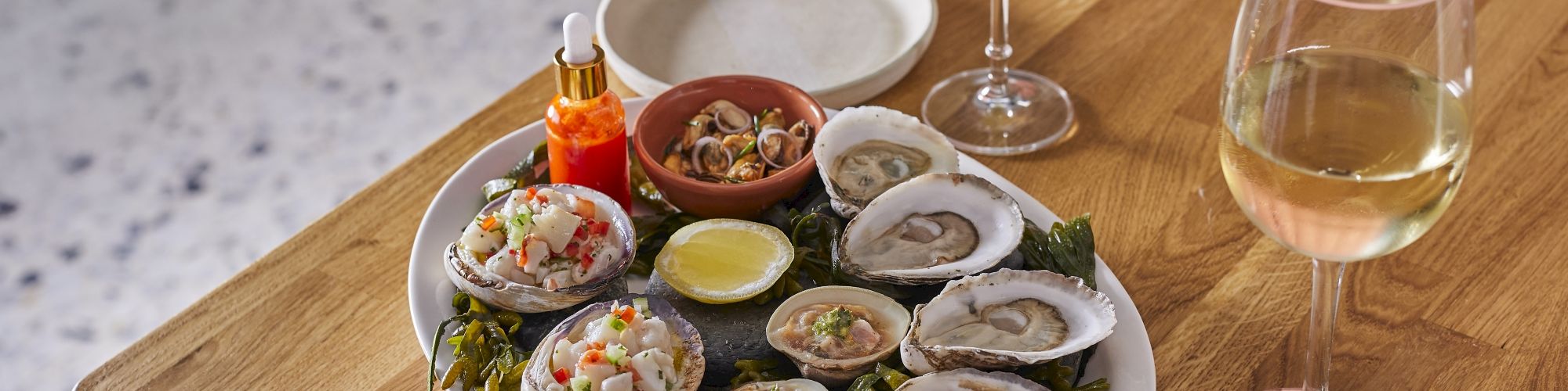 A plate of fresh oysters and clams served with hot sauce and lemon, alongside a green cocktail and a glass of white wine.