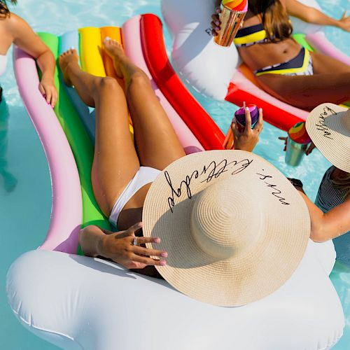 People relaxing on pool floats, wearing sun hats and swimsuits, enjoying drinks in a swimming pool.