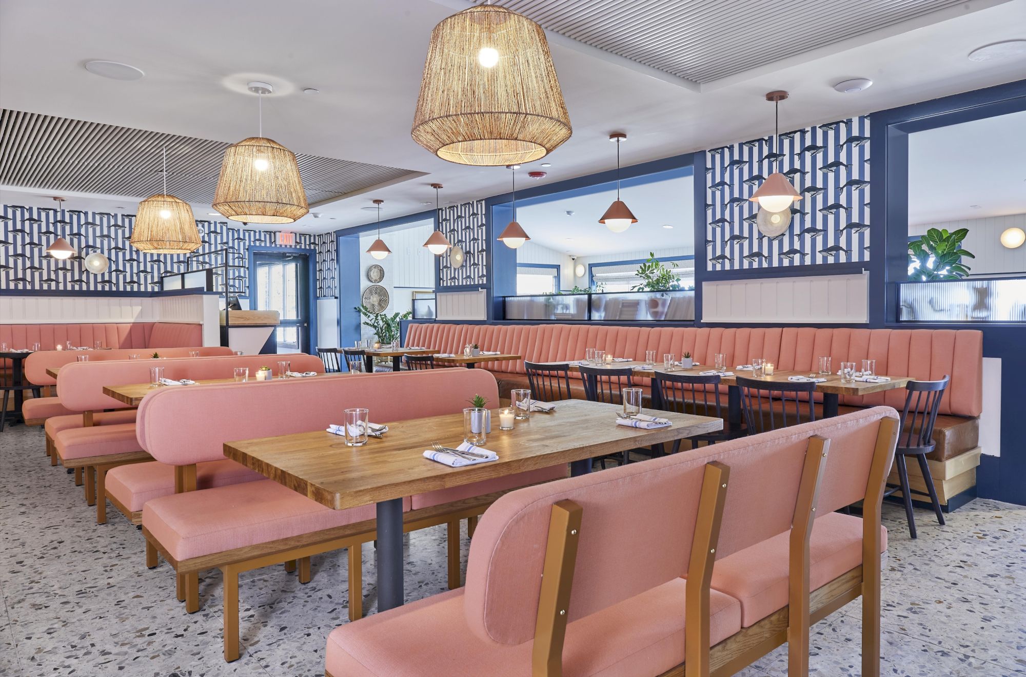 A modern restaurant interior with pink seating, wooden tables, pendant lights, and patterned walls, creating a cozy, stylish ambiance.