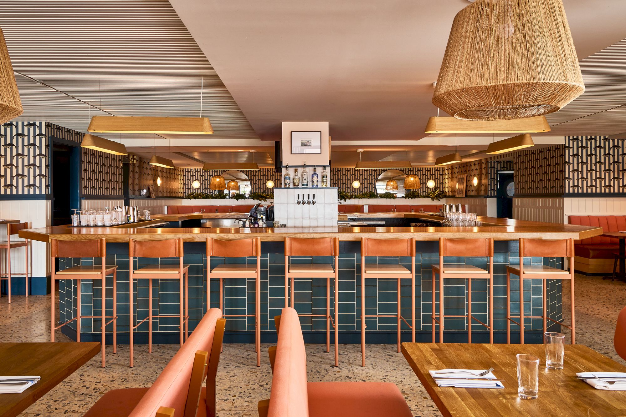 A stylish, airy bar with wooden stools, a long counter, tiled front, warm lighting, and cozy seating&mdash;ready for drinks and conversations.