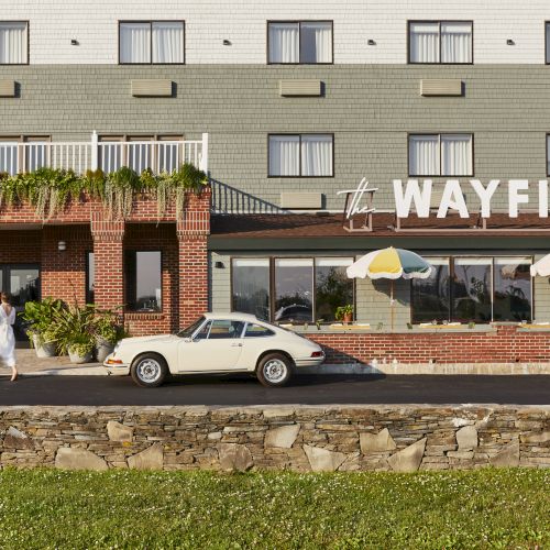 A building with "WAYFINDER" signage, a vintage car parked in front, and a person walking nearby. There's greenery and umbrellas visible.