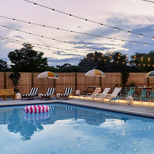 A poolside area with lounge chairs, umbrellas, hanging string lights, and a floating pool ring at dusk against a sky with scattered clouds.