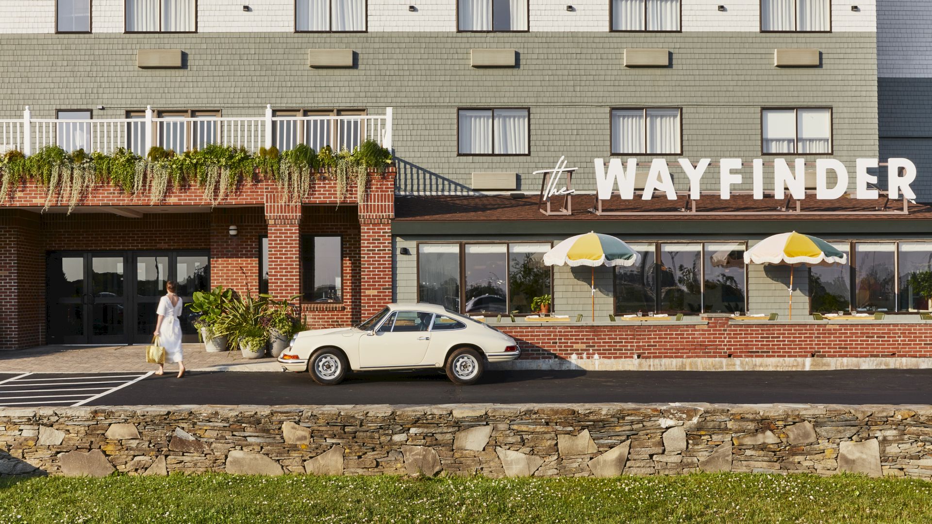 A person walks towards a vintage car parked outside a building labeled "The Wayfinder" with colorful umbrellas.