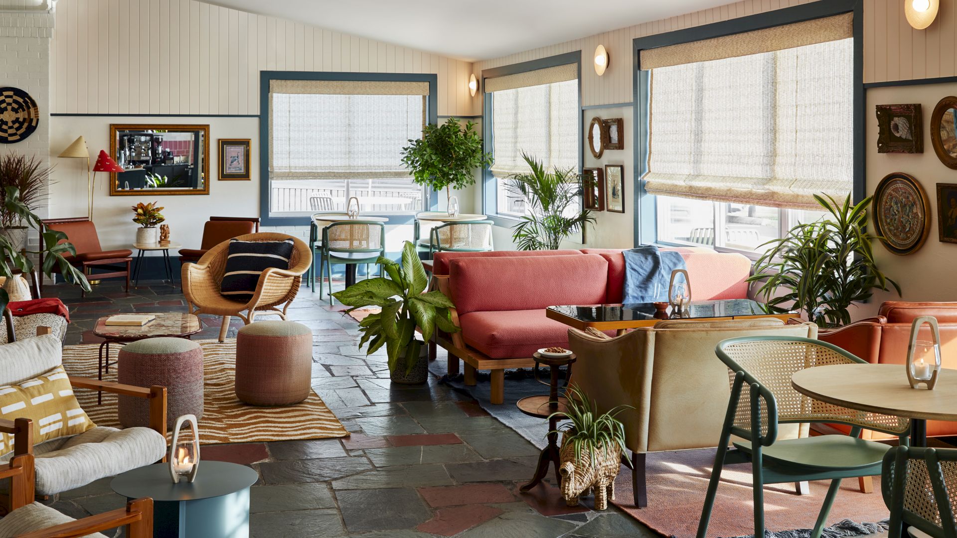 A cozy, stylish lounge with colorful sofas, chairs, plants, and framed art on the walls, filled with natural light from large windows.