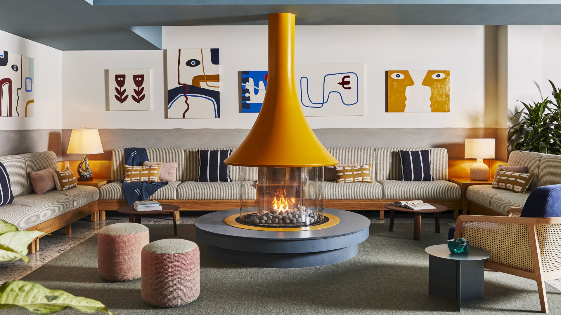 A cozy living room with a central fireplace, surrounded by modern sofas and art on the walls. Warm lighting and contemporary decor complete it.