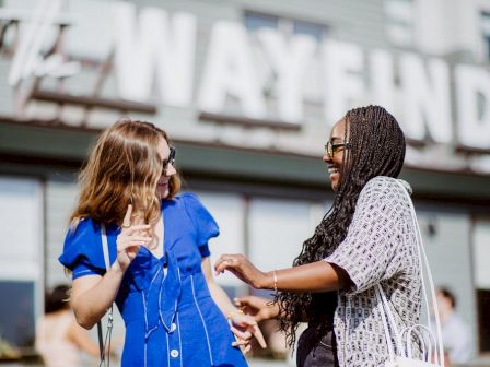 Two people are laughing and interacting in front of a building with a sign that says "The Wayfinder."