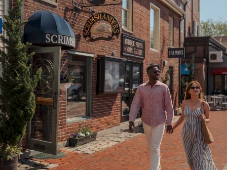 A couple walks on a brick-lined street outside shops, including a sign reading &ldquo;Scrimshanders,&rdquo; on sunny day.