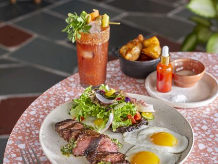 A table with steak, eggs, salad, a Bloody Mary, sauce, hot sauce, and fried potatoes on a terrazzo surface.