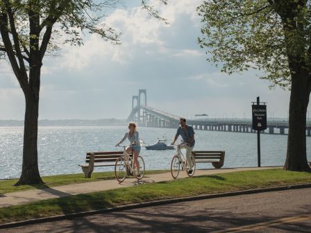 A couple rides bicycles along the waterfront in John J. Martin Memorial Park, with a bridge spanning the bay behind them.