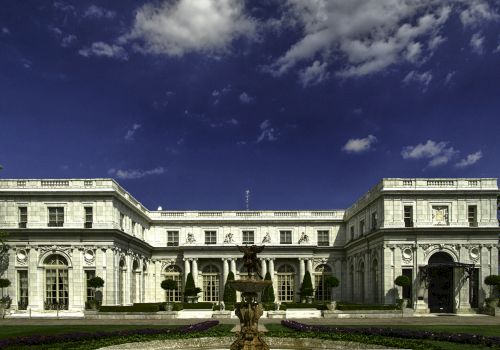 The image shows a grand neoclassical-style mansion with ornate columns and a central fountain, set against a vivid blue sky.