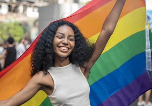A person smiling and holding a rainbow flag at an outdoor event.