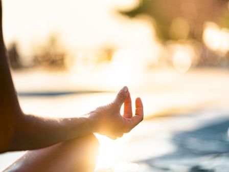 A person is meditating outside, with their hand in a relaxed position, against a blurred sunny background.
