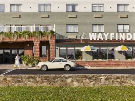 A woman walks near a vintage car parked outside "The Wayfinder" with colorful umbrellas and brick facade, under a multi-story building.