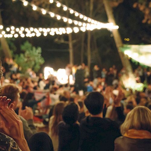 An outdoor gathering with people seated under string lights, surrounded by trees, creating a warm and festive atmosphere.