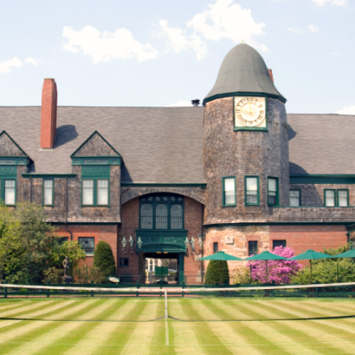 A historic building with a clock tower overlooks a grass tennis court, surrounded by greenery and flowers.