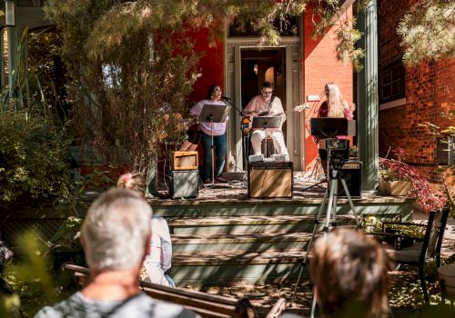 Musicians perform on a porch with instruments and microphones, while a small audience watches in a sunlit outdoor setting.