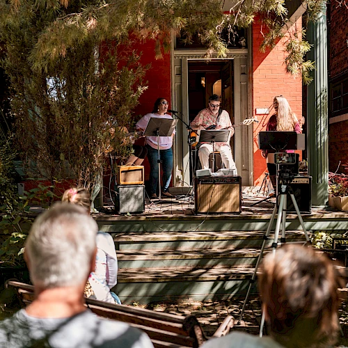 Musicians perform on a porch with instruments and microphones, while a small audience watches in a sunlit outdoor setting.
