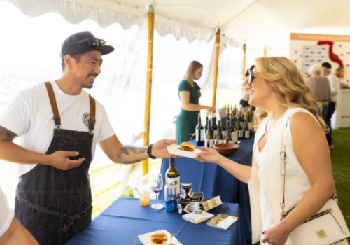 A man in an apron hands a small plate to a woman under a tent at a food and wine event.