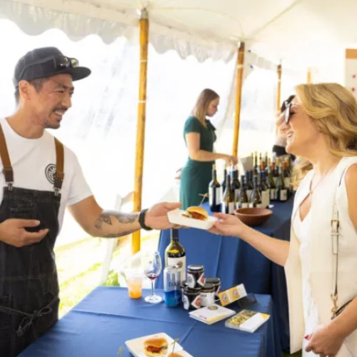 A man in an apron hands a small plate to a woman under a tent at a food and wine event.