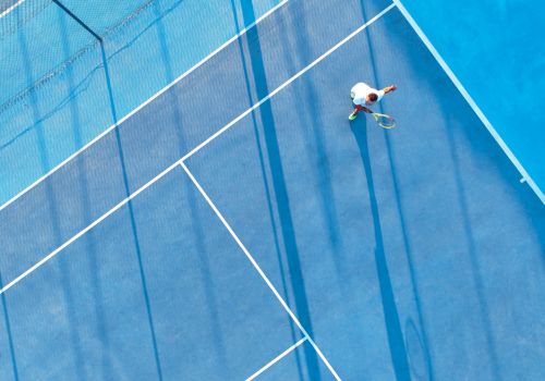 A person is playing tennis on a blue court, seen from above, with dramatic shadows stretching across the surface.