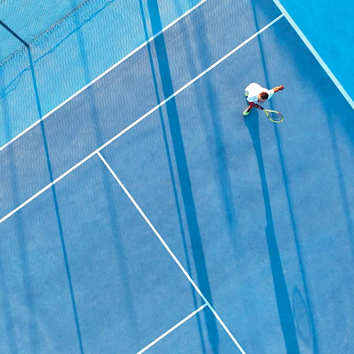 A person is playing tennis on a blue court, seen from above, with dramatic shadows stretching across the surface.