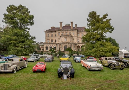 A collection of vintage cars is displayed on a lawn in front of a large historic mansion, with trees and cloudy skies in the background.