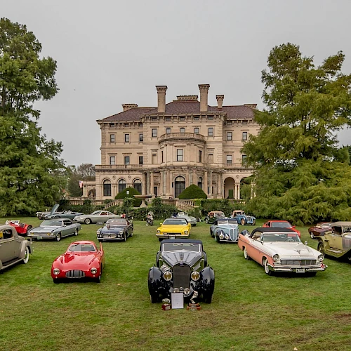 A collection of vintage cars is displayed on a lawn in front of a large historic mansion, with trees and cloudy skies in the background.
