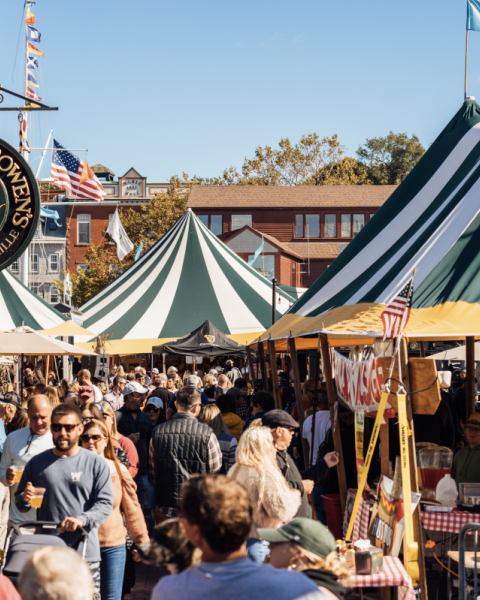A bustling outdoor market with people browsing stalls, colorful tents, and a sign for "TWENTY TWO BOWEN'S" on a nearby building catches the eye.