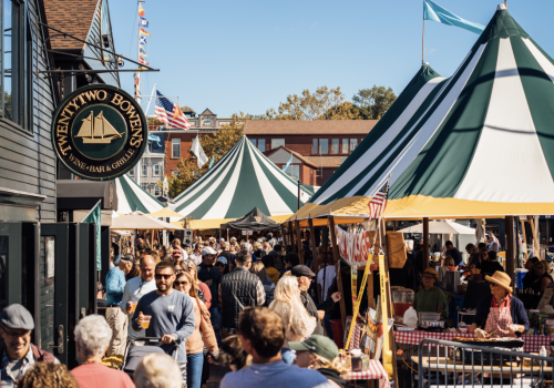 A bustling outdoor market with people browsing stalls, colorful tents, and a sign for "TWENTY TWO BOWEN'S" on a nearby building catches the eye.