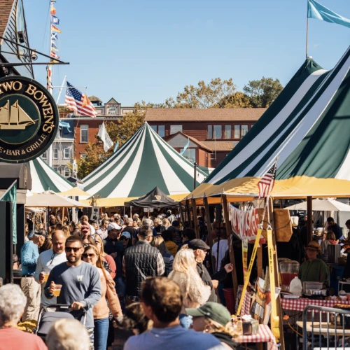 A bustling outdoor market with people browsing stalls, colorful tents, and a sign for "TWENTY TWO BOWEN'S" on a nearby building catches the eye.