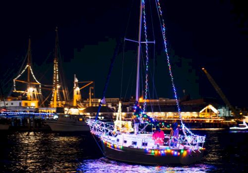 A sailboat decorated with colorful lights is floating on water at night, with other ships and a lit-up harbor in the background.