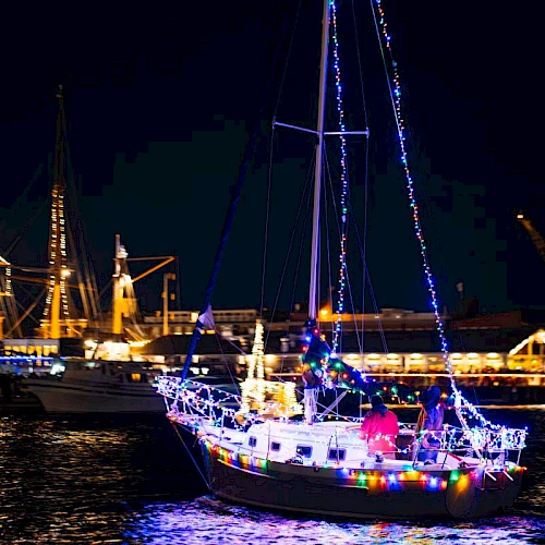 A sailboat decorated with colorful lights is floating on water at night, with other ships and a lit-up harbor in the background.