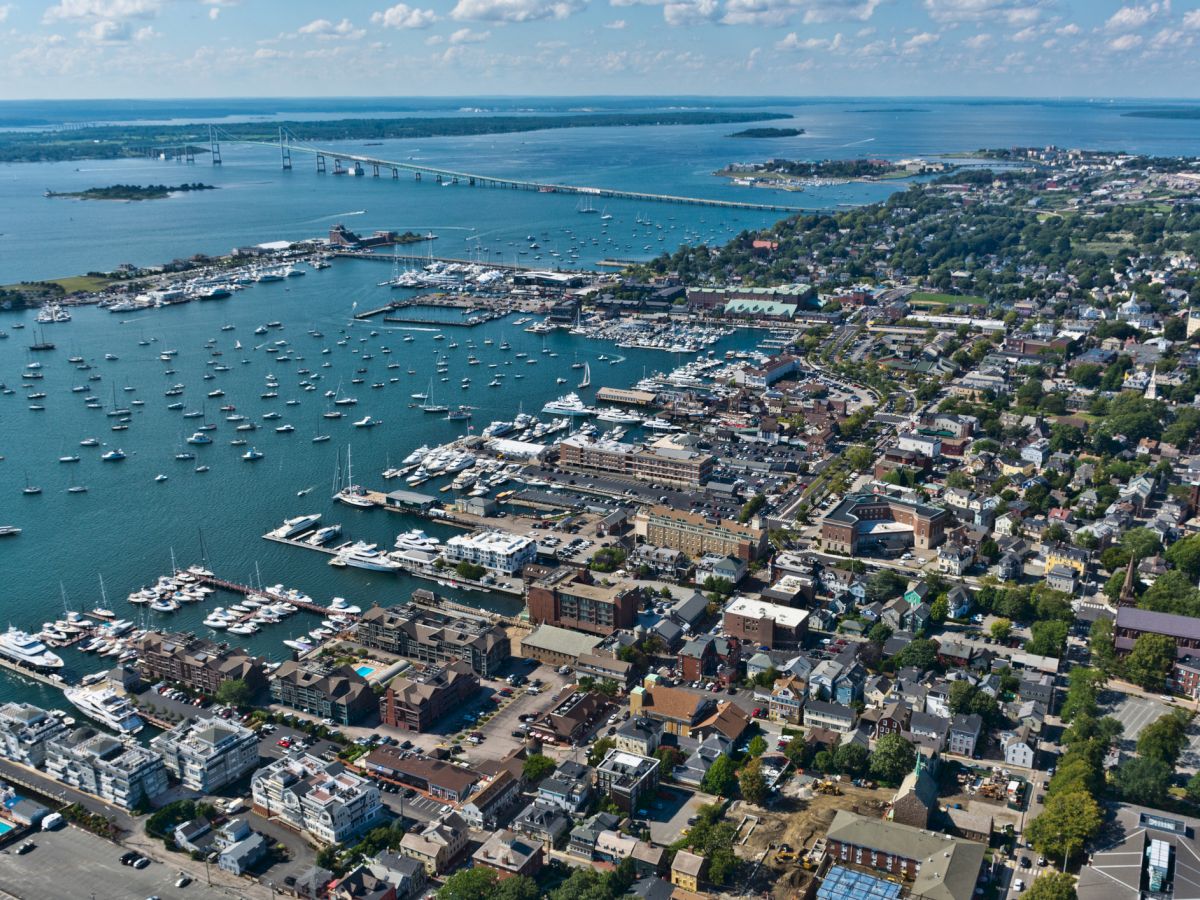 Aerial view of a coastal city with a marina full of boats, piers, and buildings near the waterfront, under a clear blue sky.