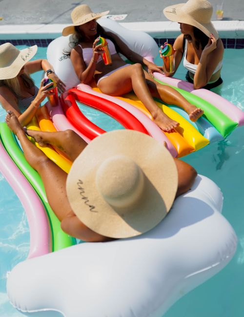 Four people in large hats relax on a colorful pool float, enjoying drinks in a sunny pool setting.