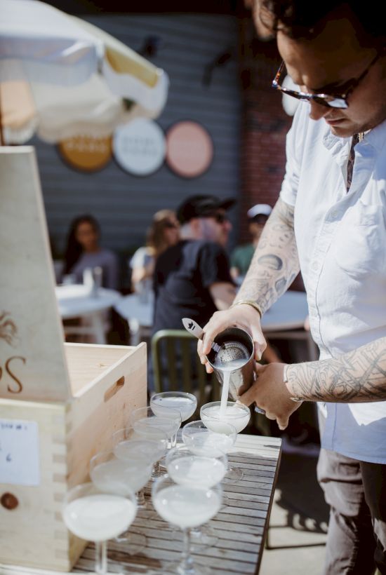 A person is pouring a drink into several glasses on a table outdoors, with people sitting in the background enjoying the event.