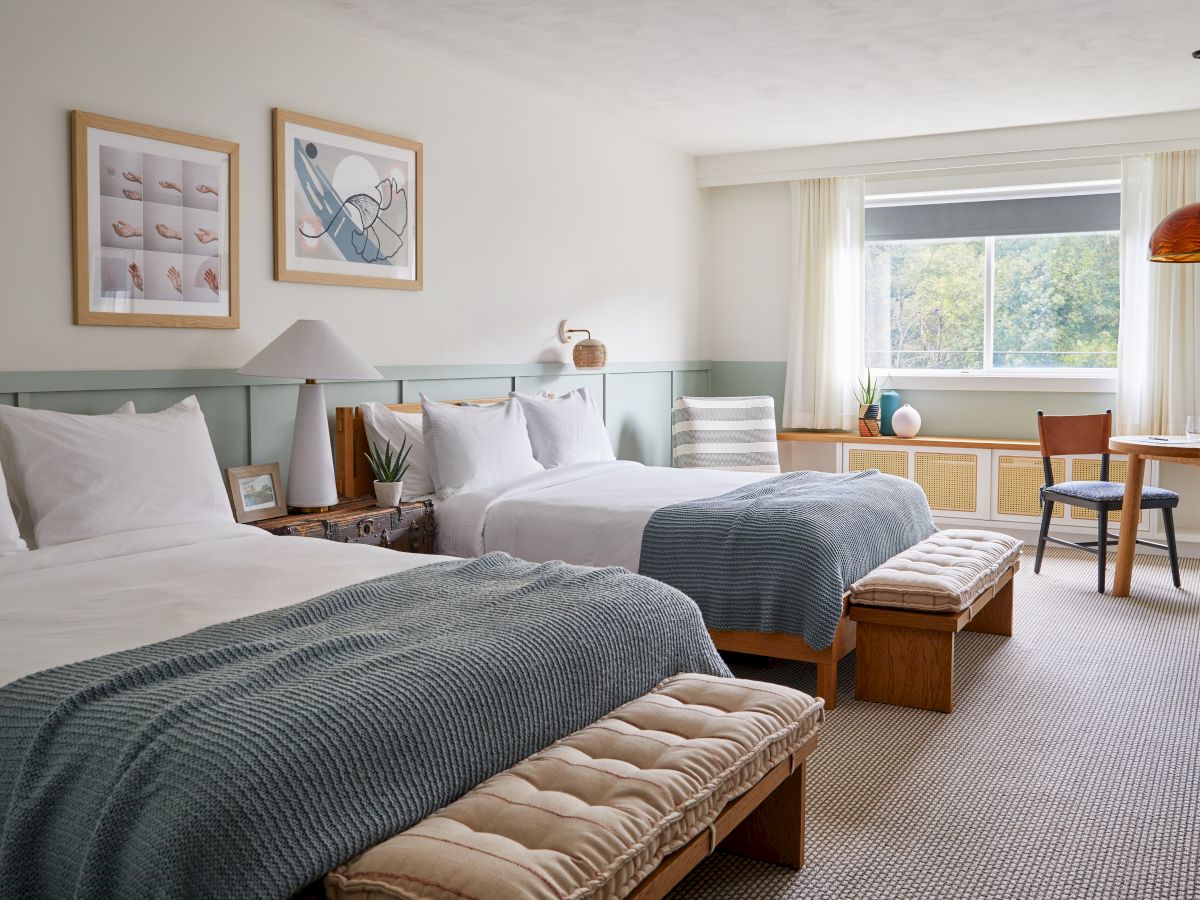 A cozy hotel room with two beds, framed art, a table by the window, and a relaxed atmosphere enhanced by warm lighting.