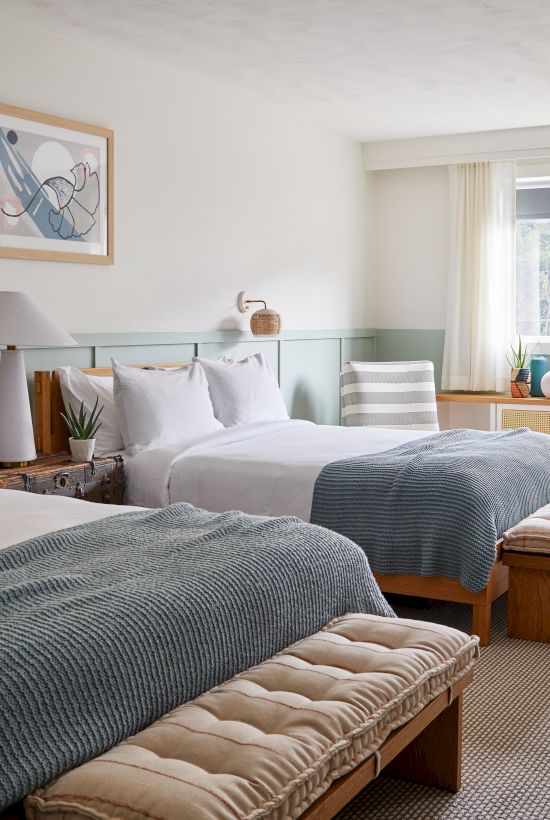 A cozy hotel room with two beds, framed art, a table by the window, and a relaxed atmosphere enhanced by warm lighting.