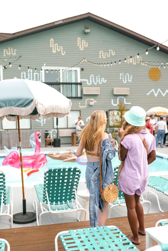 Two people stand by a pool with a pink float, surrounded by lounge chairs and umbrellas, with a decorated building in the background.
