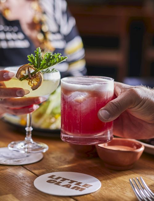 Two people clinking glasses with cocktails over a table set with food. There's a coaster labeled "Little Giant" on the wooden table.