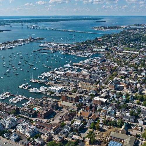 Aerial view of a coastal city with numerous boats in the water, residential and commercial buildings, and a bridge in the background.