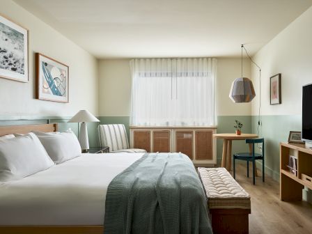 A bright modern hotel bedroom with a neatly made bed, bedside lamps, framed art, a small desk, and a TV near the window curtains.