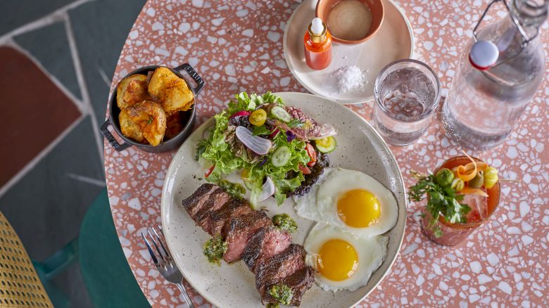 A meal with steak, two sunny-side-up eggs, salad, and roasted potatoes, accompanied by a drink, water bottle, and condiments.
