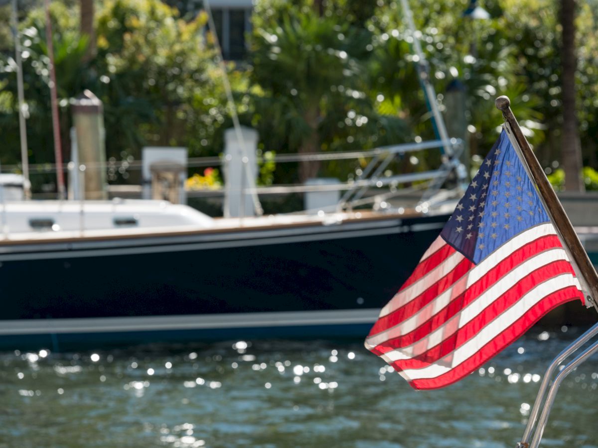 A U.S. flag waves on a boat, with another boat and lush greenery in the background, under sunny skies.