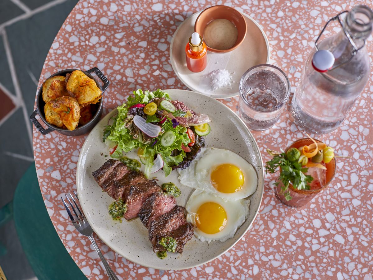 A meal with steak, eggs, and salad on a plate, roasted potatoes, drink, and condiments on a terrazzo table.