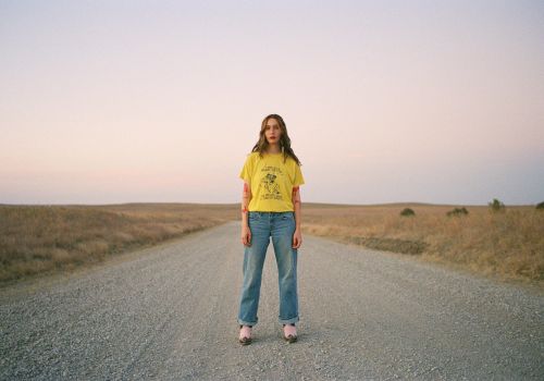 A person stands on a gravel road in an open landscape at sunset, wearing a yellow shirt and jeans.