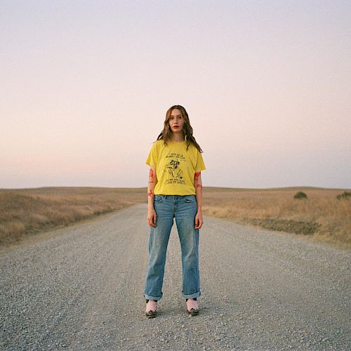 A person stands on a gravel road in an open landscape at sunset, wearing a yellow shirt and jeans.