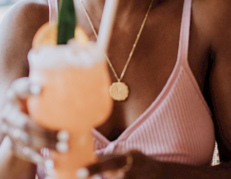 A person in a pink top holds a tropical drink with a straw and garnish, sitting at a table in a relaxed setting.
