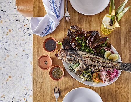 A grilled fish served with lime, vegetables, three sauces, and a drink, set on a wooden table with plates and utensils nearby.